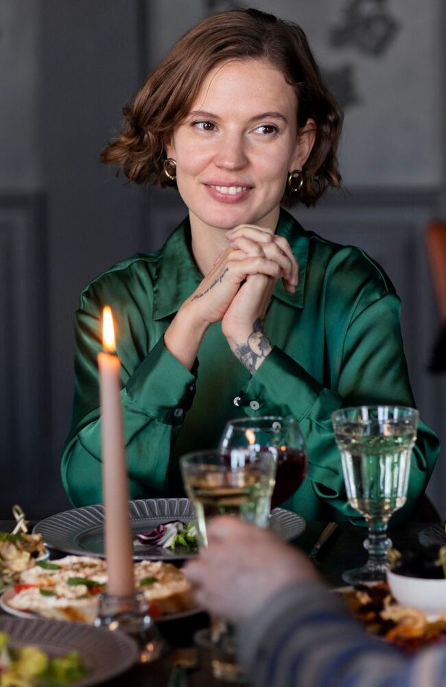 Mulher de camisa verde sorrindo à mesa de jantar com velas e convidados – a arte de receber com sofisticação